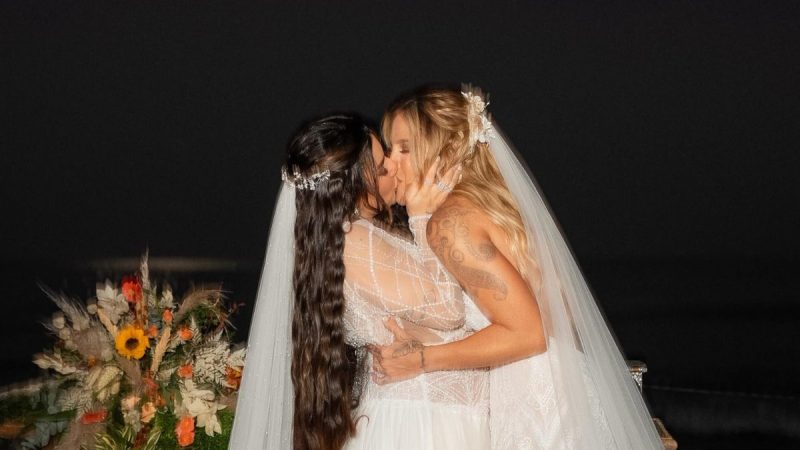 Lauana Prado e Tati Dias trocam um beijo no dia do casamento, com véu e vestido branco, em cerimônia ao ar livre com vista para o mar ao fundo.