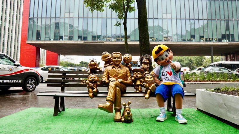 Mauricio de Sousa na Av. Paulista em homenagem aos 90 anos, em frente a um banco de bronze com esculturas do personagem Turma da Mônica, incluindo Cascão e personagens animados.