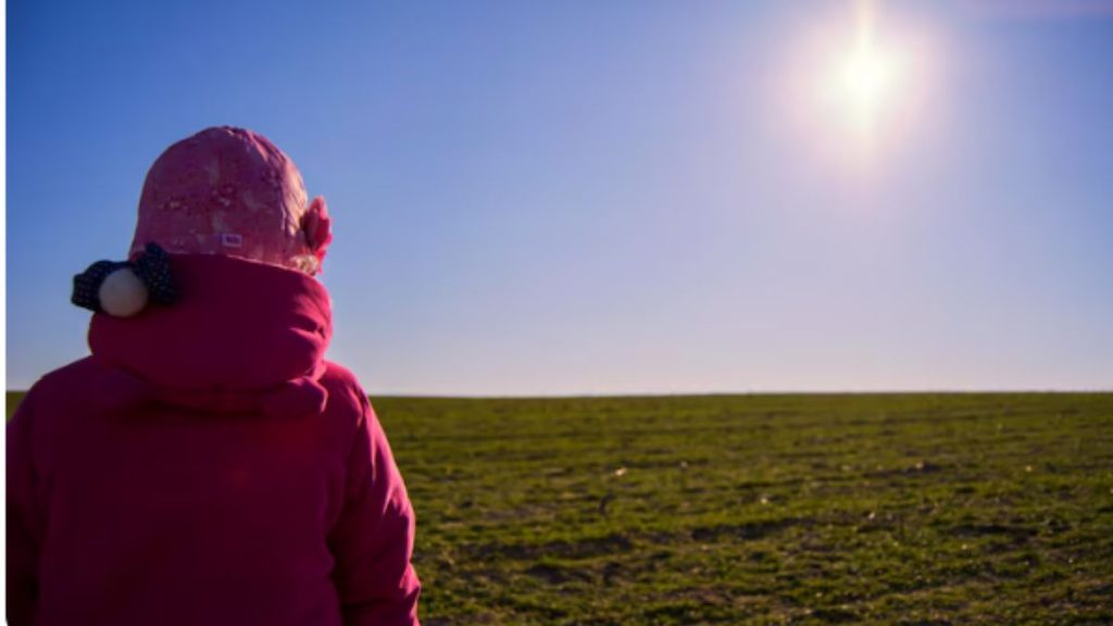 Previsão do tempo em SP: pessoa com casaco e capuz sob o sol forte no campo, com céu azul e temperaturas elevadas, indicando calor em São Paulo.