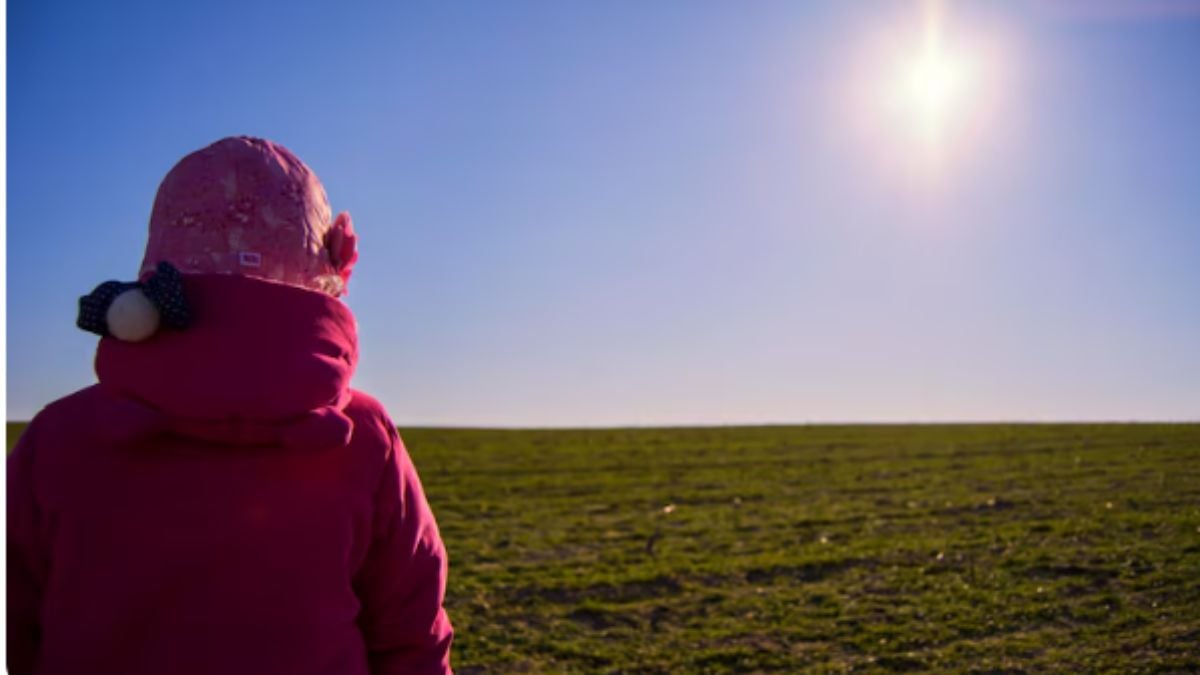 Previsão do tempo em SP: pessoa com casaco e capuz sob o sol forte no campo, com céu azul e temperaturas elevadas, indicando calor em São Paulo.