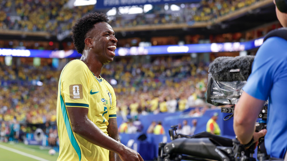 Vini Jr em campo com a camisa do Brasil, comemorando durante partida em estádio lotado, com equipe de mídia ao fundo. Destaque do Brasil na Copa.