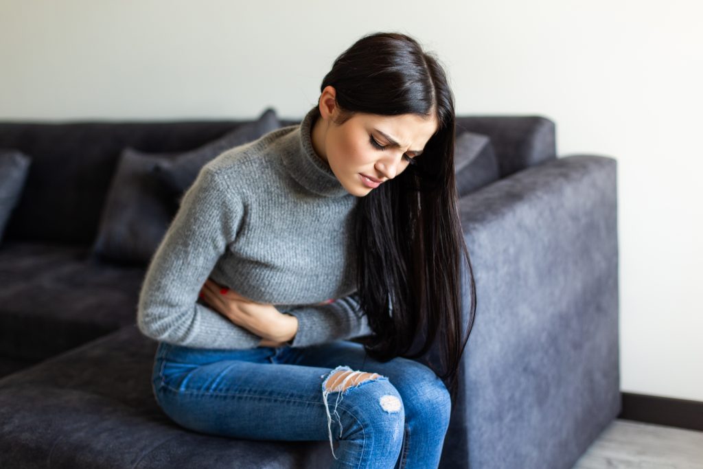 Mulher sentada em um sofá, com a mão na barriga e expressão de desconforto, simbolizando dor abdominal em campanha Julho Amarelo Limeira sobre hepatite
