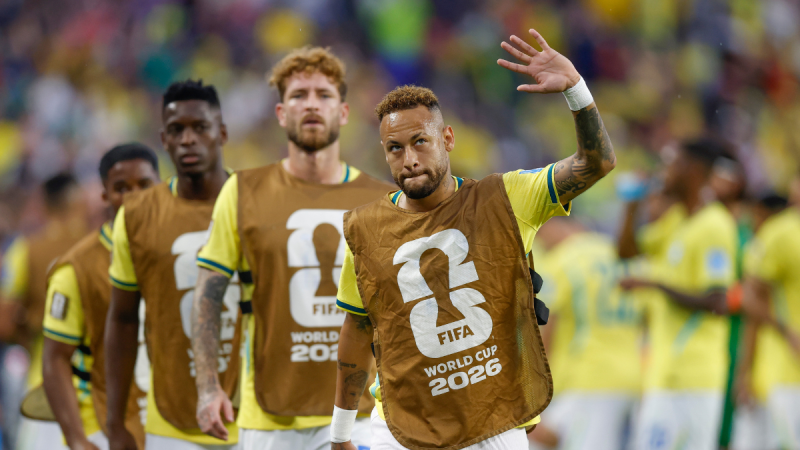 Neto Ancelotti em foto com camisa marrom e mão levantada durante transmissão esportiva, com jogadores do Brasil ao fundo em clima de discussão sobre escalação.