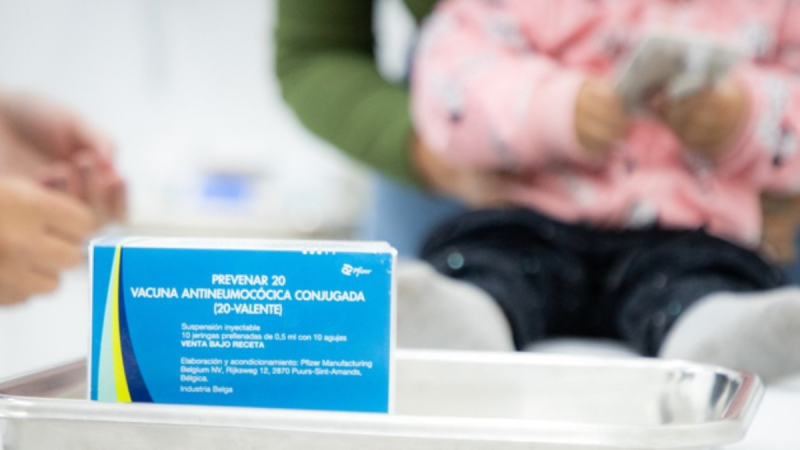 Vacinação Pneumo 20 em Sumaré: caixa da vacina Prevenar 20 na frente, com profissional ao fundo em atendimento infantil, pronta para aplicação em crianças menores de 5 anos.