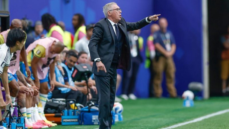 Carlo Ancelotti gesticula na linha técnica durante partida de futebol, orientando a equipe ao lado do banco de reservas, com jogadores e comissão ao fundo.