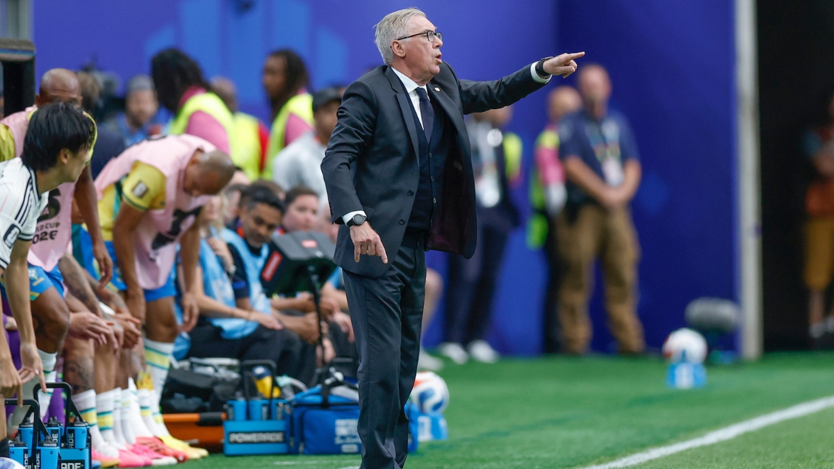 Carlo Ancelotti gesticula na linha técnica durante partida de futebol, orientando a equipe ao lado do banco de reservas, com jogadores e comissão ao fundo.