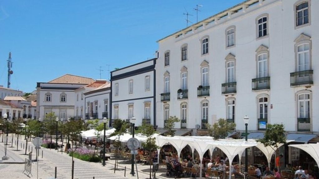 Verão em Portugal com edifícios históricos brancos e terraço de café na rua, mesas cobertas e pessoas ao ar livre sob céu azul