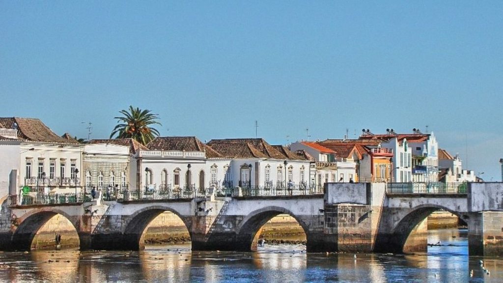 Verão em Portugal: rio com reflexos sob um conjunto de arcos de pedra de uma ponte histórica, ao fundo casas e edifícios tradicionais da cidade sob céu azul claro