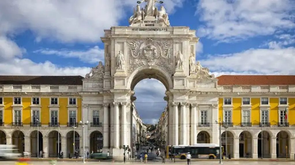 Verão em Portugal: vista da majestosa Arco da Rua Augusta em Lisboa, com edifícios amarelos e brancos ao fundo, céu azul e nuvens, ideal para turismo.