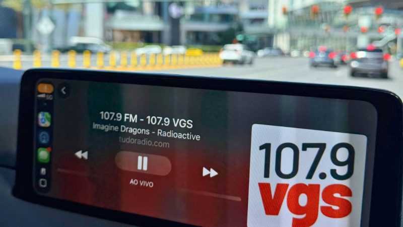 Rádio nos carros em uma tela de multimídia automotiva exibindo 107,9 VGS ao vivo com a frequência FM e informações da programação, em trânsito