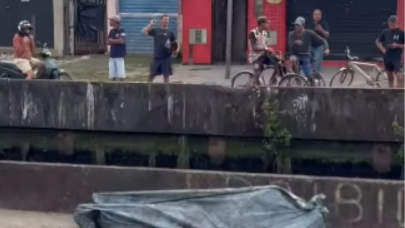 Homem morto em canal de Praia Grande: corpo submerso é avistado e coberto por lona sobre a água em área portuária, com equipes e curiosos ao fundo