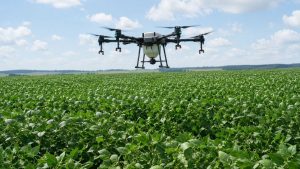 Drones agrícolas sobrevoando uma plantação verde ao ar livre, com a estrutura de voo e câmeras visíveis, destacando a automação no manejo da lavoura.