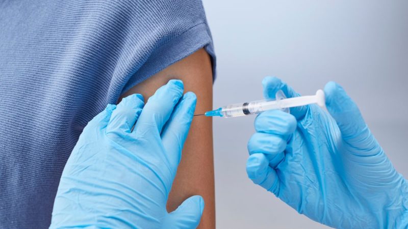 Profissional de saúde prepara e aplica vacina contra HPV em um braço, com luvas e seringa visíveis; contexto de prorrogação da vacinação até dezembro