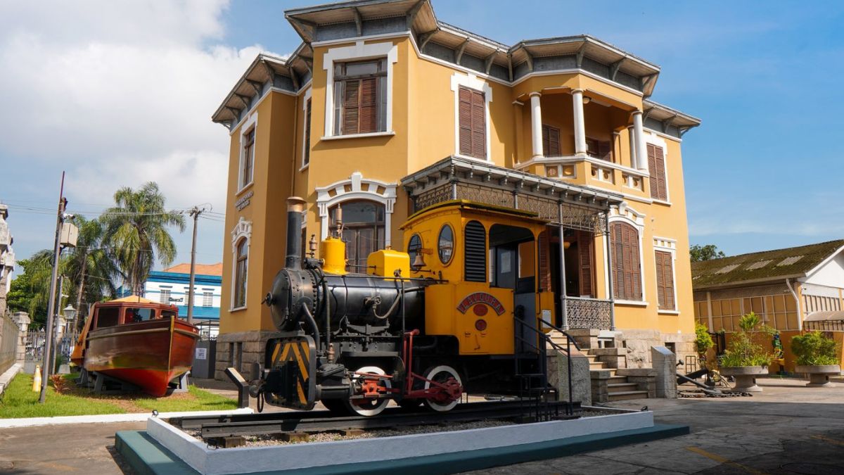 Museu do Porto de Santos: locomotiva a vapor em exposição em frente ao prédio amarelo histórico, com trilho e céu azul ao fundo. Programação gratuita nas férias de julho.