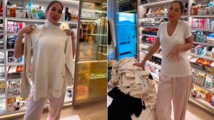 Maíra Cardi em loja de alto padrão, escolhendo roupas em provador com araras e vitrines ao fundo, usando look casual com camiseta clara e calça listrada e segurando peças de vestuário.