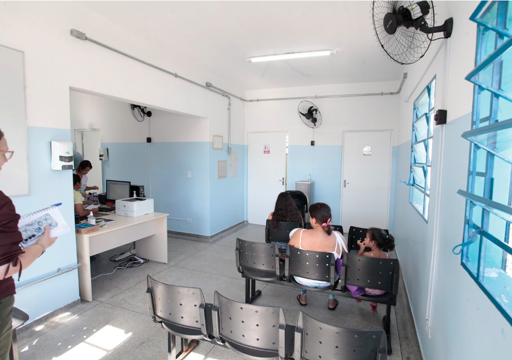 Sala de atendimento em uma UBS, com cadeiras em fila e profissionais trabalhando ao fundo, mostrando estrutura interna de unidade de saúde para assistência diurna