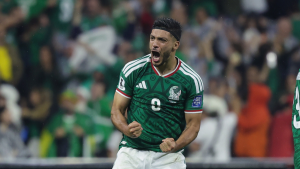 Jogador da seleção do México em campo, com camisa verde, comemorando durante partida contra o Equador; torcida ao fundo e clima de classificação às oitavas da Copa.