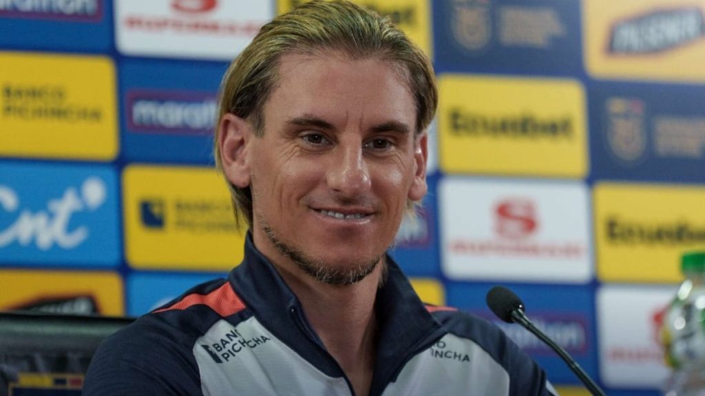 Beccacece durante coletiva de imprensa, sorrindo ao lado de microfones, em fundo com painéis de patrocinadores e clubes, em contexto de jogo