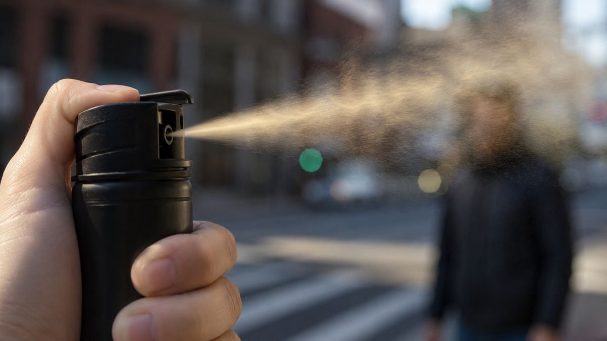 Mão segurando um spray de pimenta acionado, com jato de gás saindo do bico em ambiente urbano desfocado, simbolizando defesa pessoal e aprovação no Senado.