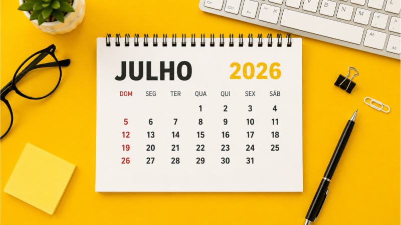 Calendário de julho 2026 sobre mesa amarela com a capa “JULHO 2026”, marcando dias da semana, com caneta, clipes, óculos e bloco de notas ao redor.