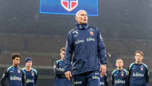 Técnico da Noruega em pé no campo durante partida, usando agasalho azul com escudo do país; ao fundo, outros jogadores da equipe observam no gramado.