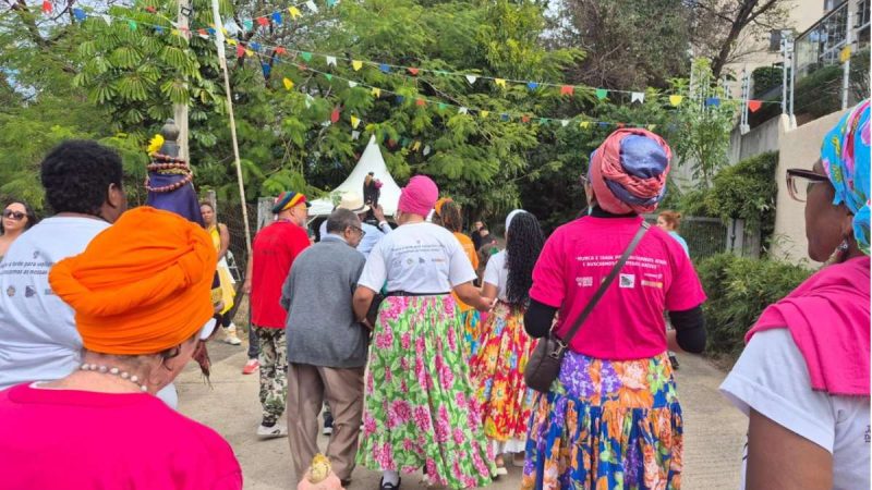 Pessoas caminham em uma rua decorada com bandeirolas coloridas durante o Arraial Afro Julino, em Campinas, com participantes usando roupas e camisetas do evento