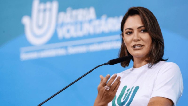 Michelle Bolsonaro em evento no microfone, usando camiseta branca com símbolo verde, em cenário azul ao fundo, posando durante pronunciamento.