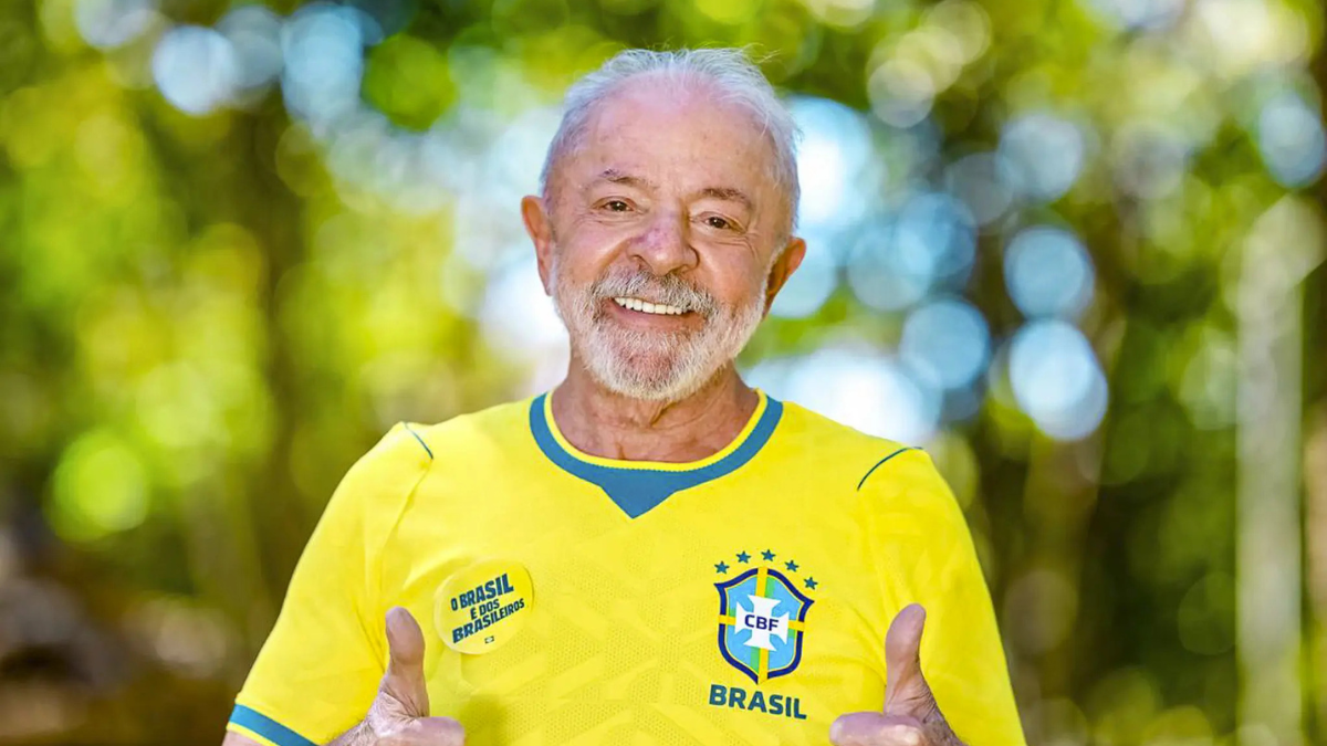 Lula, em foto sorridente, veste camisa amarela da seleção brasileira e faz gesto com as mãos em meio a fundo desfocado verde e azul. Lula pede a Ancelotti vitória do Brasil contra a Noruega.