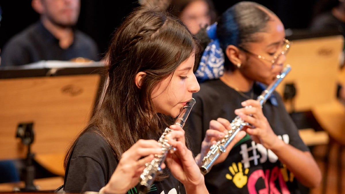 Músicos da Orquestra Popular do GURI tocando flauta transversal e flauta em apresentação ao vivo, com instrumentos e músicos ao fundo em palco.