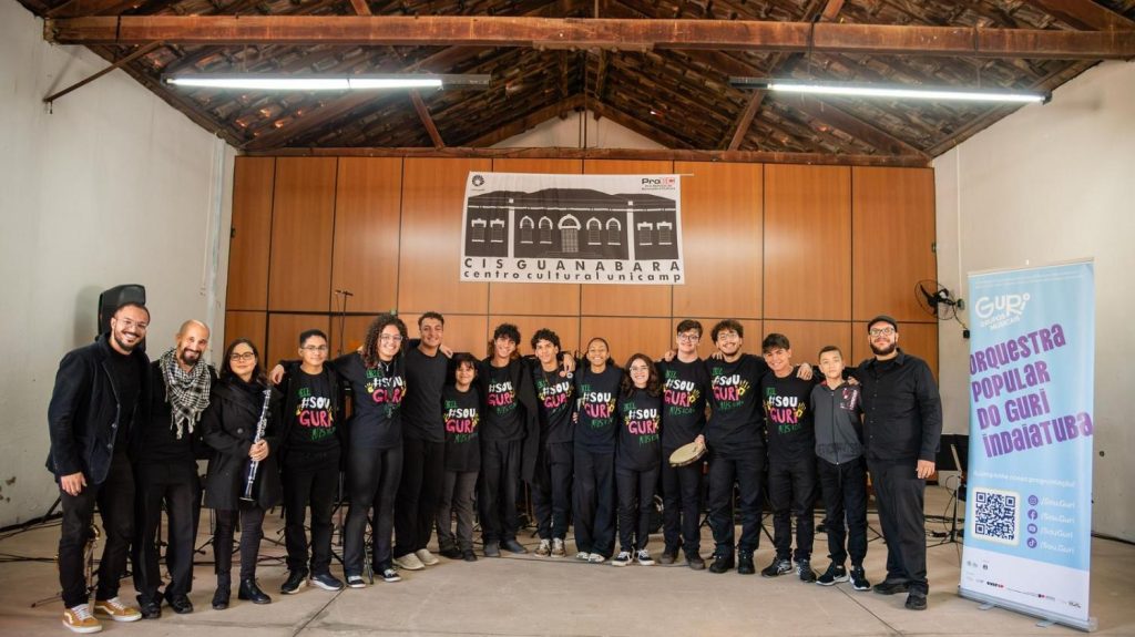 Foto de grupo da Orquestra Popular do GURI em um palco, com integrantes usando camisetas do projeto e um banner institucional ao fundo, em ambiente de apresentação e ensaio.