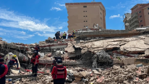 Atualizações terremotos na Venezuela: equipes de resgate entre escombros após o colapso de prédios, com pessoas usando equipamentos de proteção em meio a destroços e entulho sob céu azul.