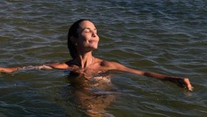 Ivete Sangalo aparece em foto na água, tomando banho de mar ao pôr do sol, com expressão serena e cabelo preso, enquanto flutua nas ondas.