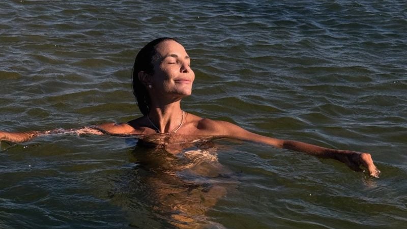 Ivete Sangalo aparece em foto na água, tomando banho de mar ao pôr do sol, com expressão serena e cabelo preso, enquanto flutua nas ondas.