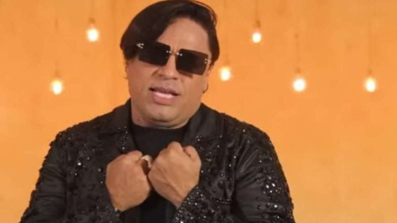 Neto Araújo, cantor da Collo de Menina e ex-integrante dos Cavaleiros do Forró, aparece em foto com óculos escuros, jaqueta preta e iluminação quente ao fundo