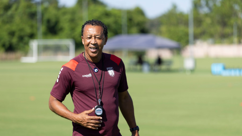 Técnico de Cabo Verde em treino no campo de futebol, com camisa marrom e apito/credencial no pescoço, gesticulando com confiança durante preparação para enfrentar a Argentina.