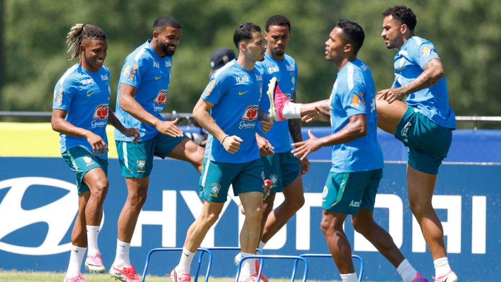 Jogadores da seleção brasileira em treino ao ar livre, com Camisas azuis e faixa de aquecimento, comemorando e se cumprimentando em campo com estrutura ao fundo; Danilo Santos e Martinelli são opções para o confronto com a Noruega.