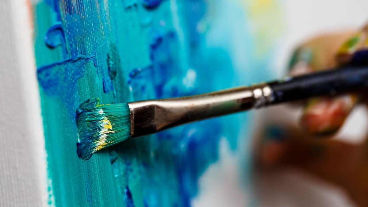 Pincel com cerdas pintando uma tela azul em pintura artística, destacando a divulgação das Oficinas Culturais em Indaiatuba com inscrições a partir de 13 de julho.