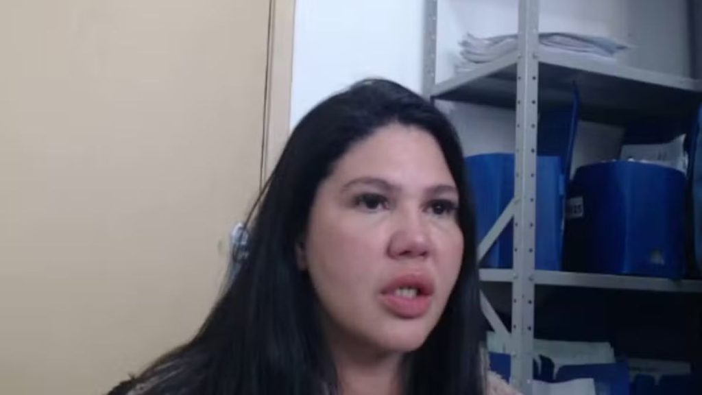Eveline dos Santos Gonçalves aparece em vídeo com cabelo escuro e expressão séria, em frente a uma estrutura metálica com caixas azuis, em contexto de indiciamento pela Polícia Civil.