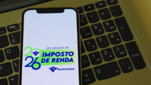 Imagem mostra um celular com a tela da Declaração de Imposto de Renda 2024/2025 ao lado de um teclado de computador, contexto de consulta do cashback do Imposto de Renda.