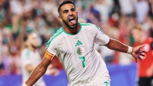 Mahrez, jogador da seleção da Argélia, aparece em campo comemorando com as mãos abertas e expressão de satisfação durante partida da Copa do Mundo, em imagem que acompanha anúncio de despedida após eliminação.