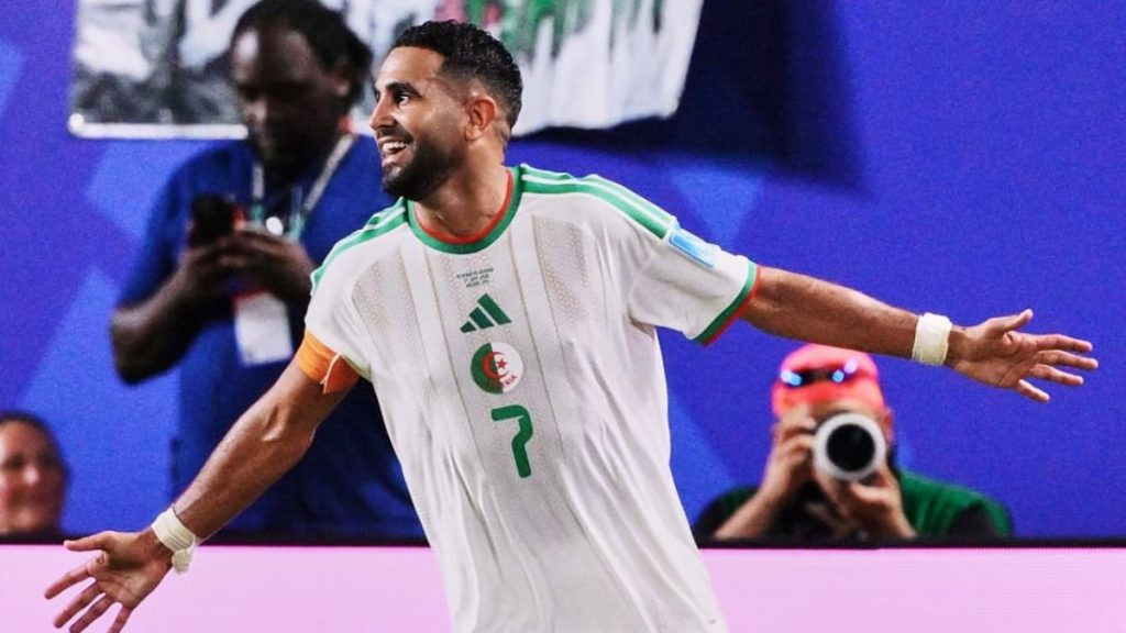 Mahrez comemora em campo, usando a camisa da Argélia branca com detalhes verdes, braços abertos e expressão de celebração durante uma partida.