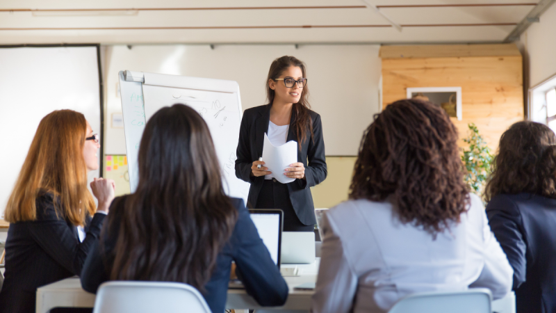 Instrutora em sala de aula para turma sentada, apontando conteúdo em quadro branco e distribuindo material, cenário de capacitação com inscrições para cursos gratuitos na Casa da Mulher de Santos