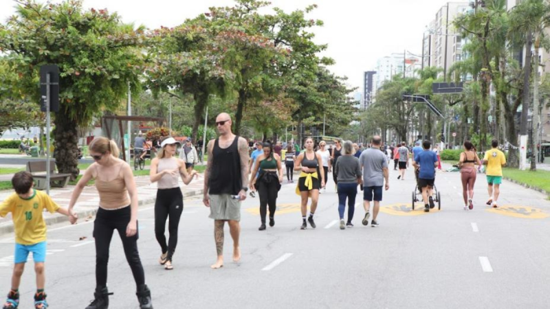 Vem Pra Orla Santos: pessoas caminhando e trânsito urbano na orla, com faixas e veículos ao fundo, indicando alterações de itinerários e circulação no domingo