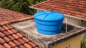 Caixa d’água gratuita Sabesp azul instalada sobre laje com telhado de telhas cerâmicas ao fundo, em área residencial da Baixada Santista. Informativo sobre quem tem direito.