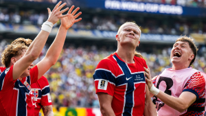 Haaland Brasil em campo comemorando vitória, com o atacante sorrindo e levantando as mãos, enquanto jogadores ao redor celebram na torcida ao fundo.