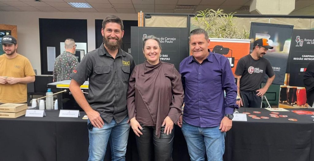 Equipe em estande de feira gastronômica ao lado de banners da Rota da Cerveja de São Paulo, com quatro pessoas posando em frente a mesas e materiais de divulgação.