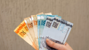 Mão segura vários cédulas de dinheiro brasileiro com o valor de 20 reais, representando o prazo do quinto dia útil do mês de julho para o pagamento do salário em julho 2026.