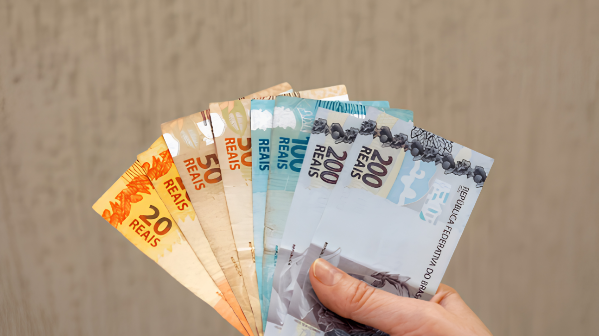 Mão segura vários cédulas de dinheiro brasileiro com o valor de 20 reais, representando o prazo do quinto dia útil do mês de julho para o pagamento do salário em julho 2026.