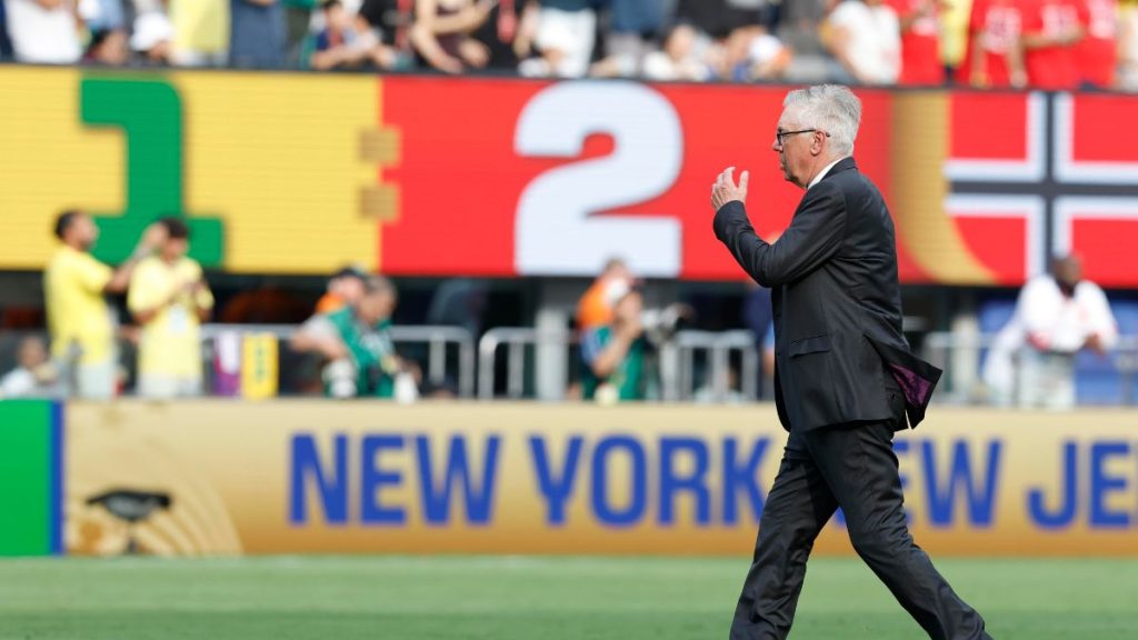 Técnico Carlo Ancelotti, da Seleção Brasileira, ao lado do campo durante partida em estádio, com placas ao fundo e bandeiras, enquanto se discute a eliminação do Brasil. Público e bancos visíveis
