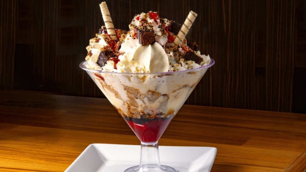 Dia do Chocolate com taça de sobremesa gelada de chocolate e chantilly, coberta com pedaços de barra, calda vermelha e confeitos coloridos sobre mesa de madeira.